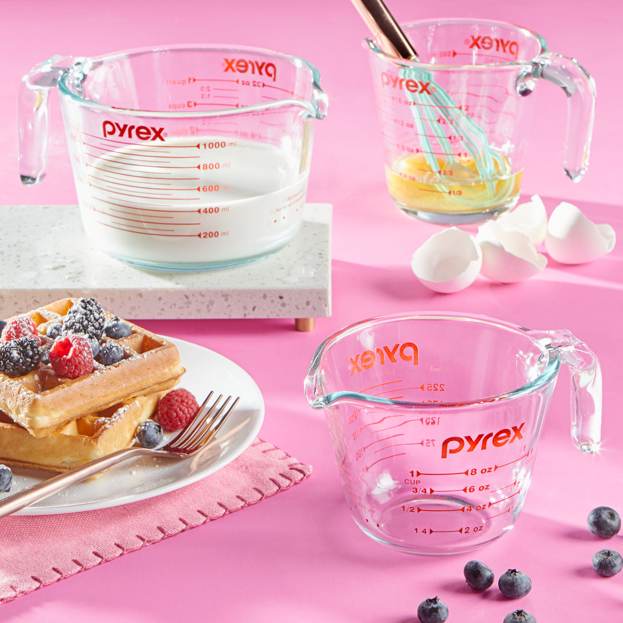 Pyrex measuring cups with waffles, berries, and a whisk on a pink background