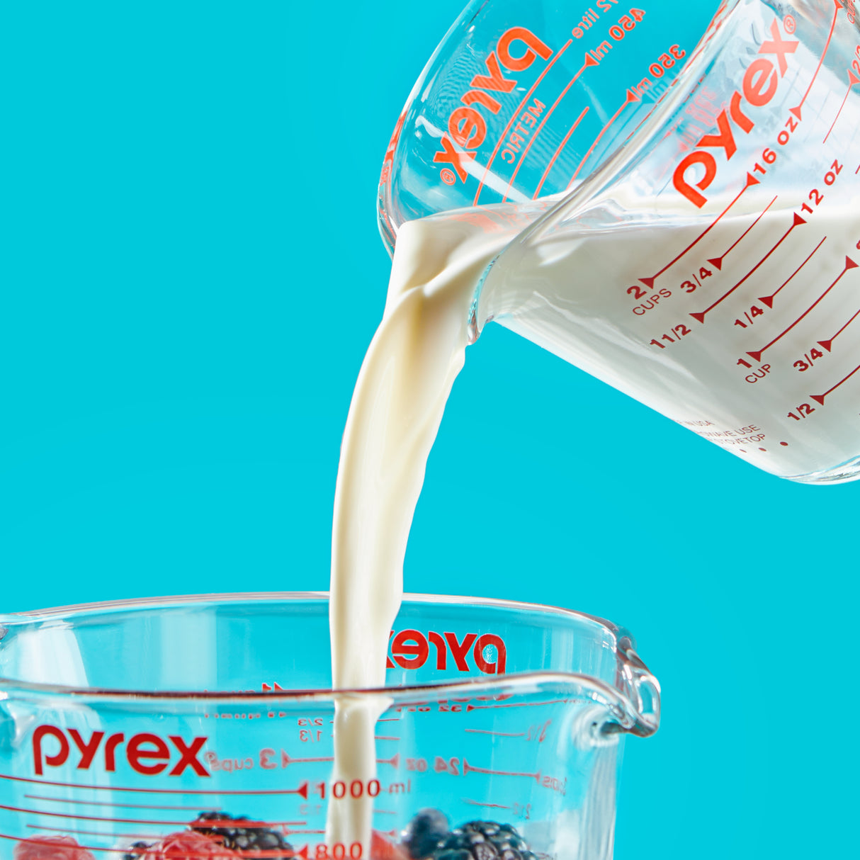 Pyrex measuring cup pouring milk into another Pyrex container against a blue background