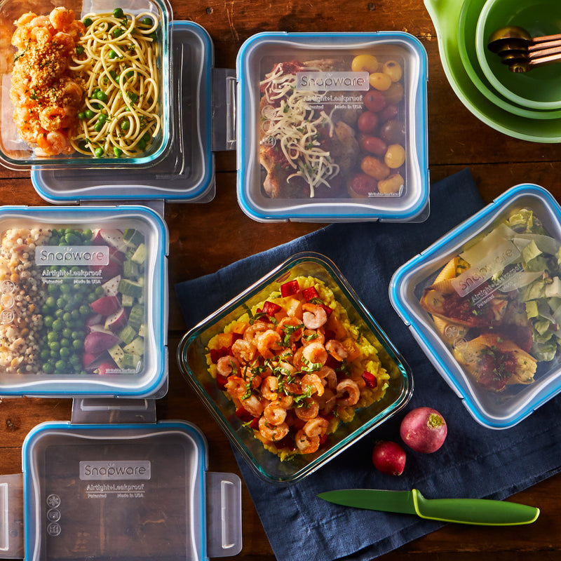 Glass meal prep containers with pasta, shrimp, vegetables, and salads on a wooden table.