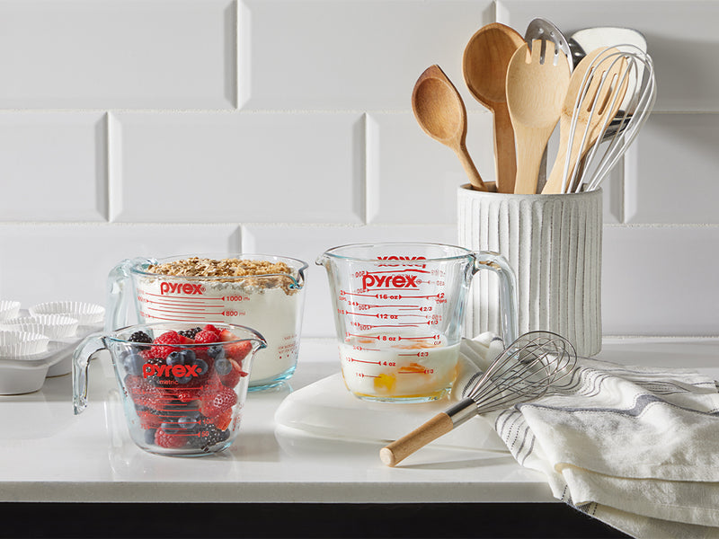 Pyrex measuring cups with berries and eggs on a kitchen counter, surrounded by wooden spoons and a whisk.