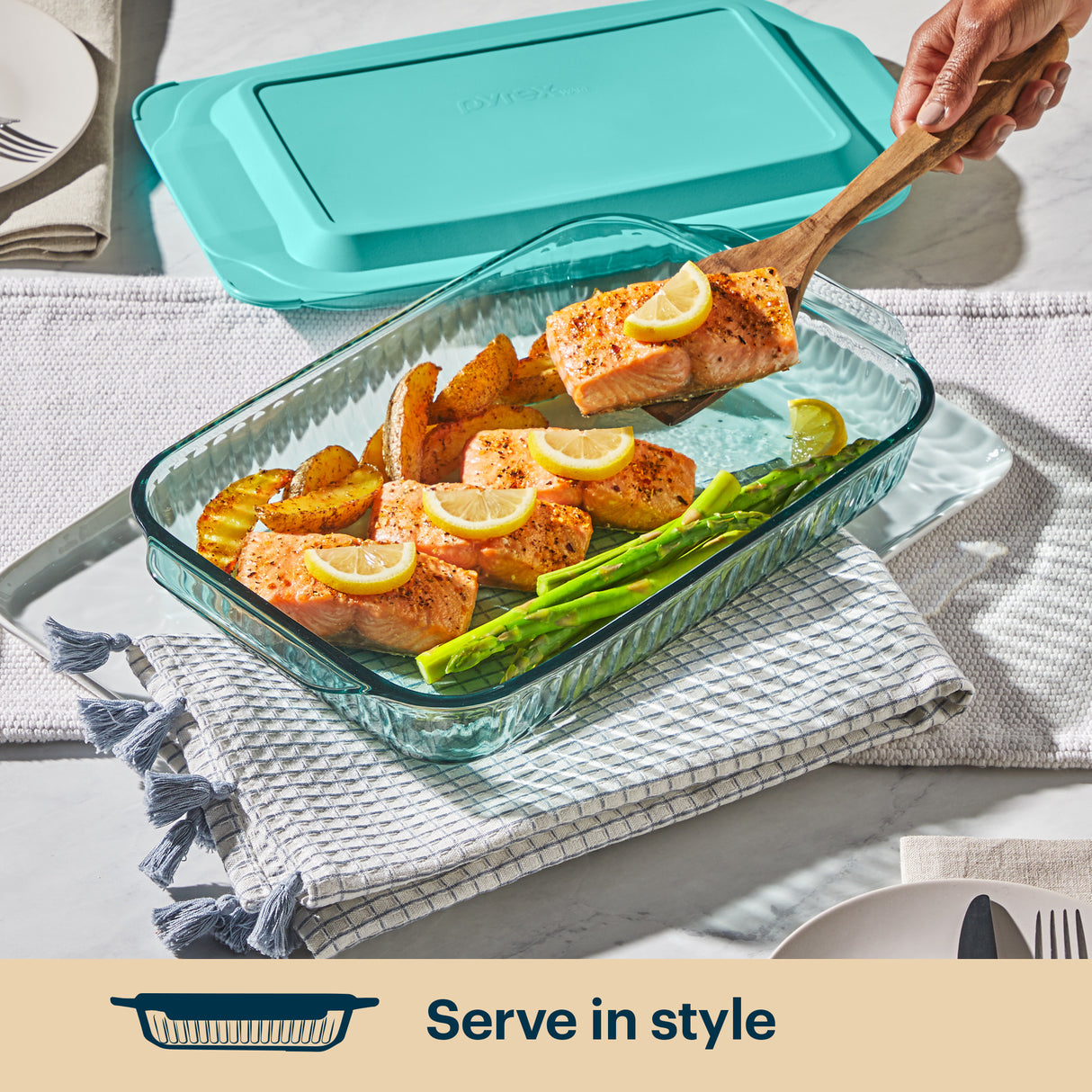 Glass baking dish with salmon, asparagus, and lemon slices on a table with a teal lid.