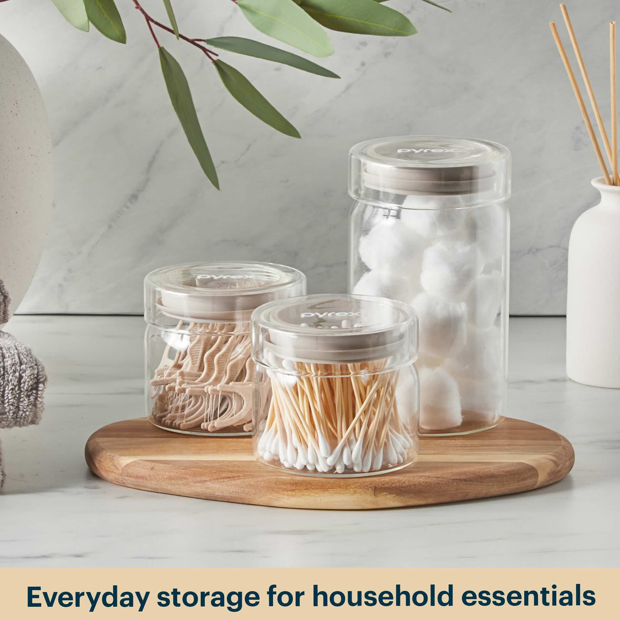 Clear jars with lids containing household essentials on a wooden tray with a neutral background.