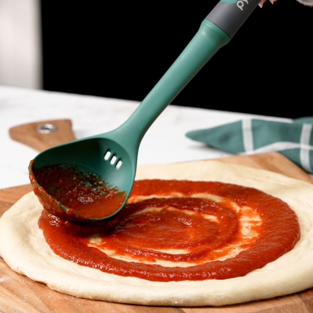 Green ladle spreading tomato sauce on pizza dough with a blurred background