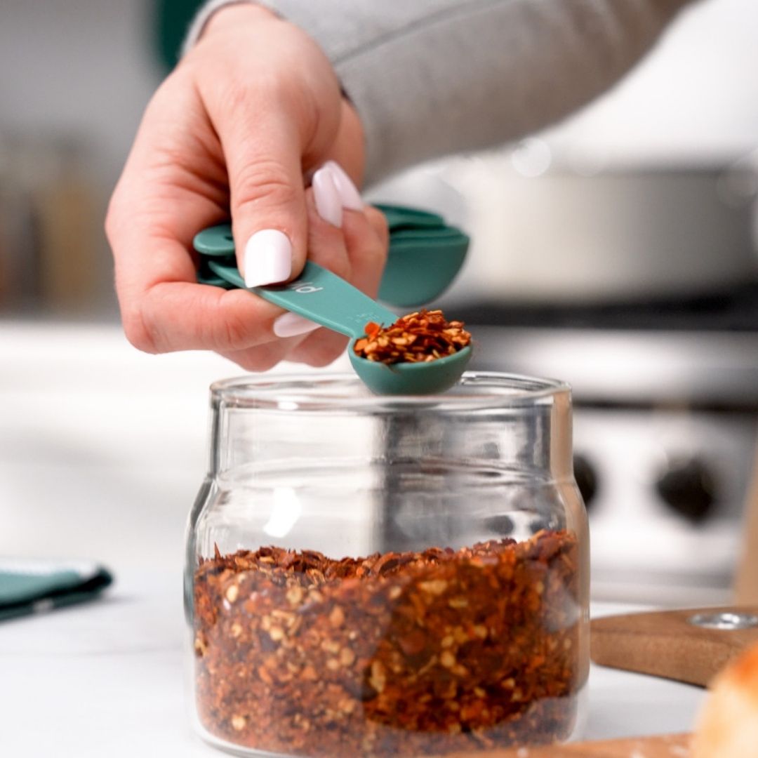 Person measuring spices with a green spoon into a glass jar on a kitchen counter.
