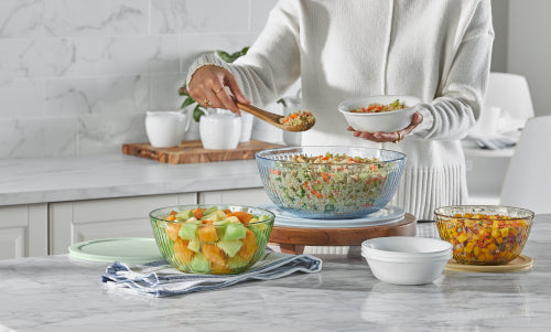 Person serving food from glass mixing bowls filled with salad, fruit, and salsa on a kitchen counter.