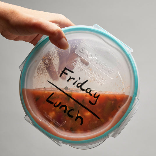 Hand holding a round Snapware container labeled 'Friday Lunch' filled with soup.