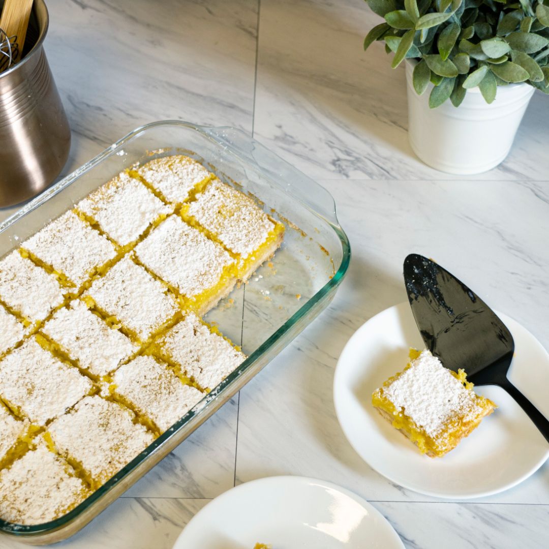 Glass dish of sliced lemon bars with one on a plate on a marble countertop.
