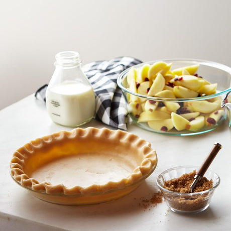Pie plate with dough and brown sugar next to it with apples and milk on the background