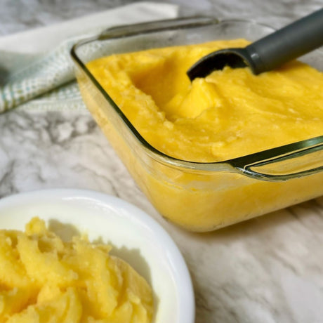 Glass container with yellow puree and a spoon, next to a bowl of puree on a marble surface.