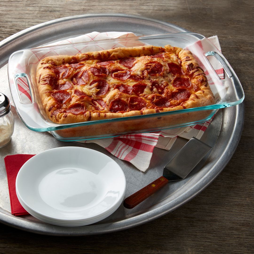 Glass dish with pepperoni pizza on a metal tray with plates and a spatula.