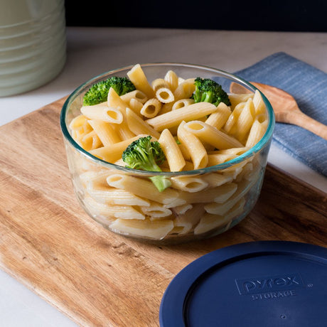 Glass bowl of pasta with broccoli on a wooden cutting board, featuring a Pyrex lid.