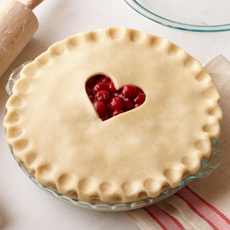 Pie with a heart-shaped cutout on top, filled with red fruit, on a white surface.