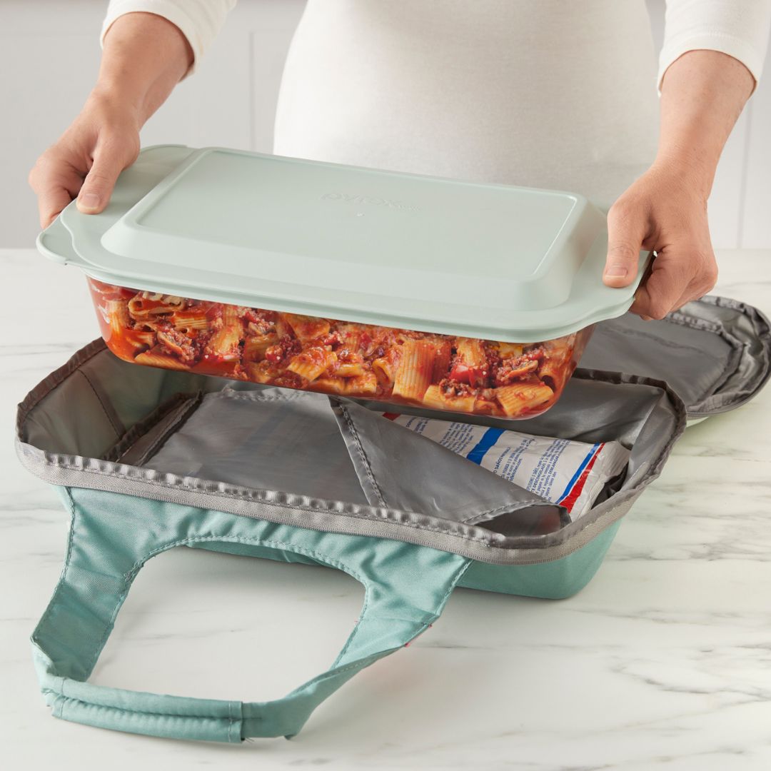 Person holding a baking dish with a lid over a portable oven on a light-colored surface.