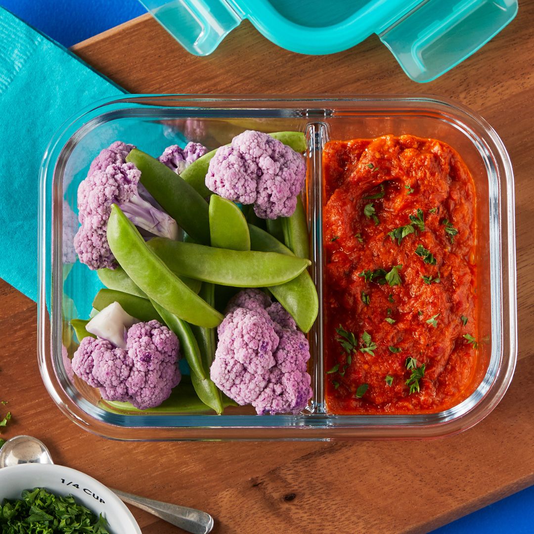 Glass lunch container with compartments of roasted red pepper dip and mixed vegetables on a wooden surface.