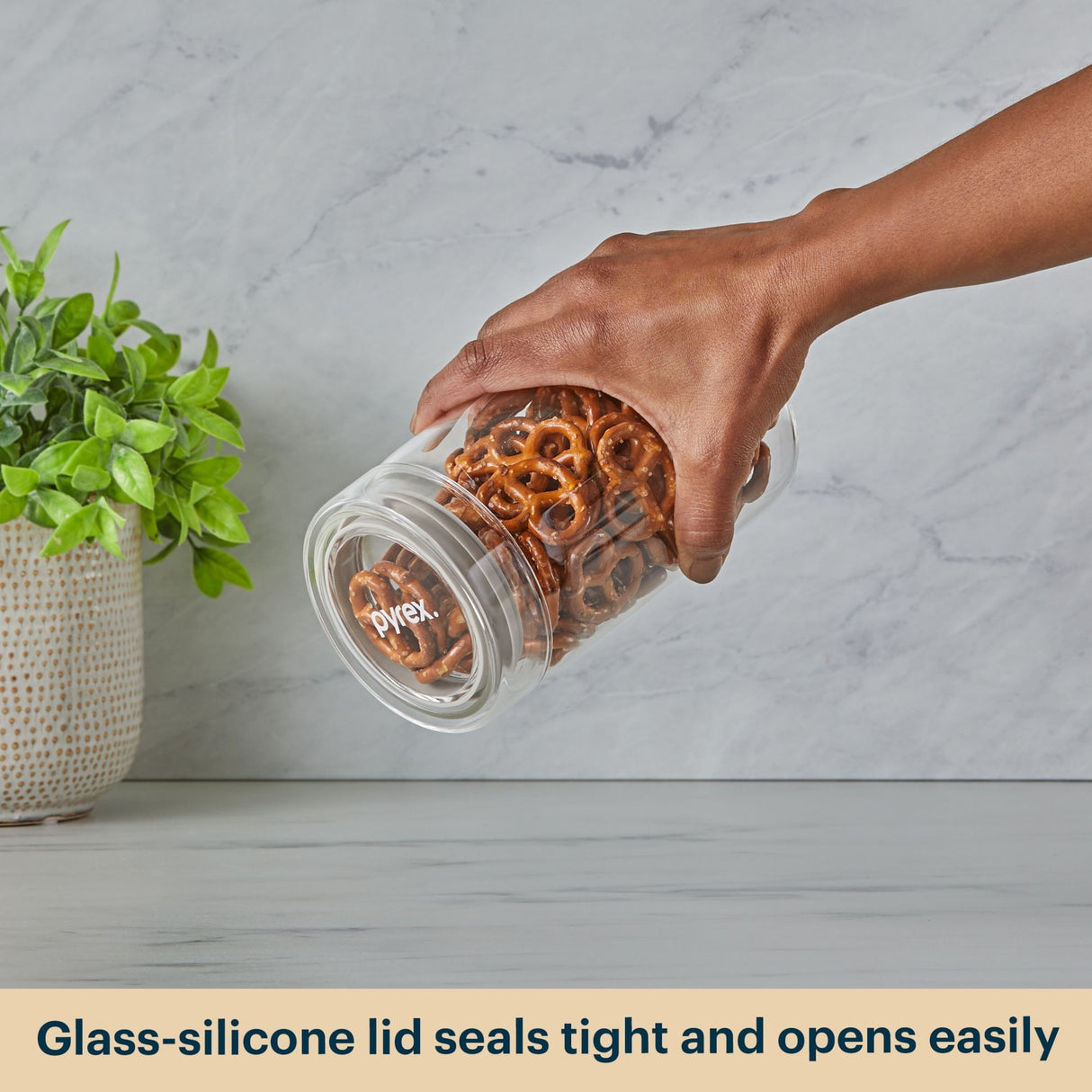 Hand holding a Pyrex jar with pretzels on a light gray surface with a plant in the background.