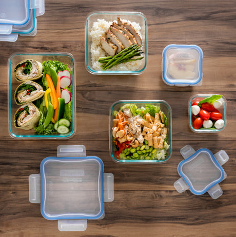 Stack of clear plastic food storage containers with blue locking lids, filled with fresh vegetables and food.