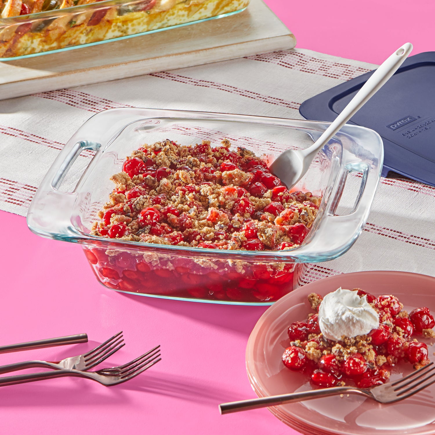 Glass baking dish with cherry cobbler on a pink surface with a white cloth underneath.