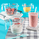 Pyrex measuring cups with fruits, a smoothie, and a spatula on a checkered tablecloth.