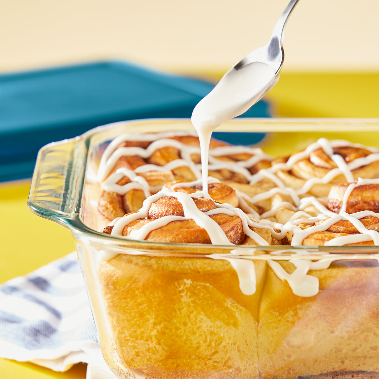 Casserole dish with cinnamon rolls being drizzled with white icing on a yellow surface.