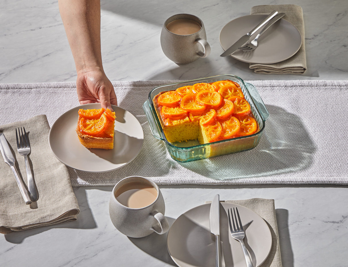 Person serving a slice of orange cake from a glass dish on a dining table with coffee cups and plates.