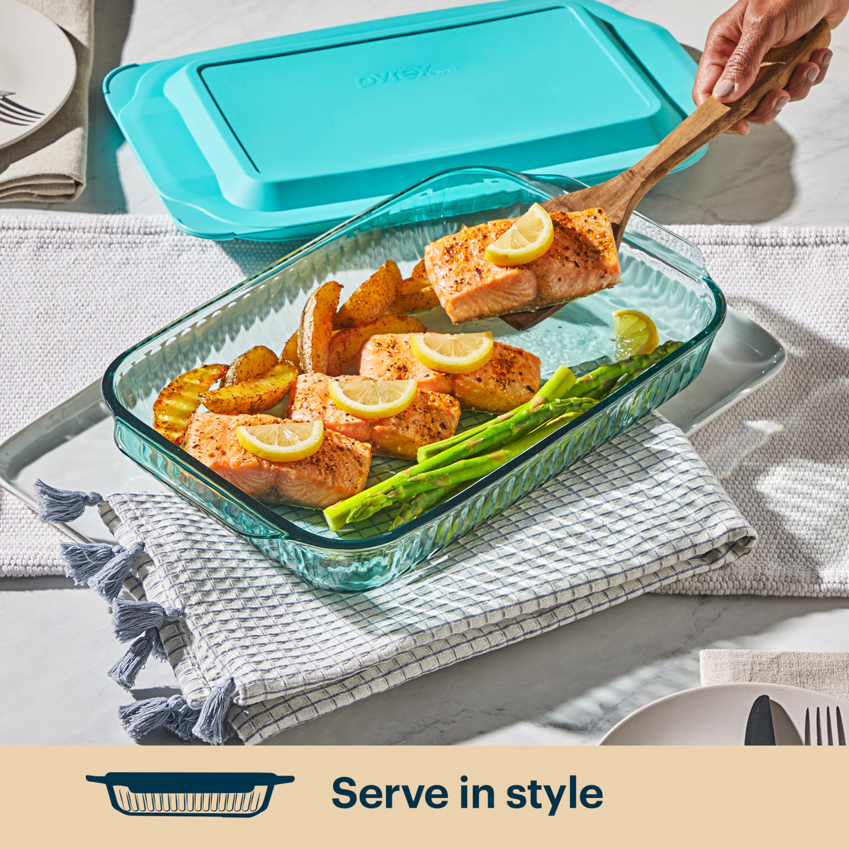 Glass baking dish with salmon, asparagus, and lemon slices on a table with a blue lid.