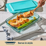 Glass baking dish with salmon, asparagus, and lemon slices on a table with a blue lid.