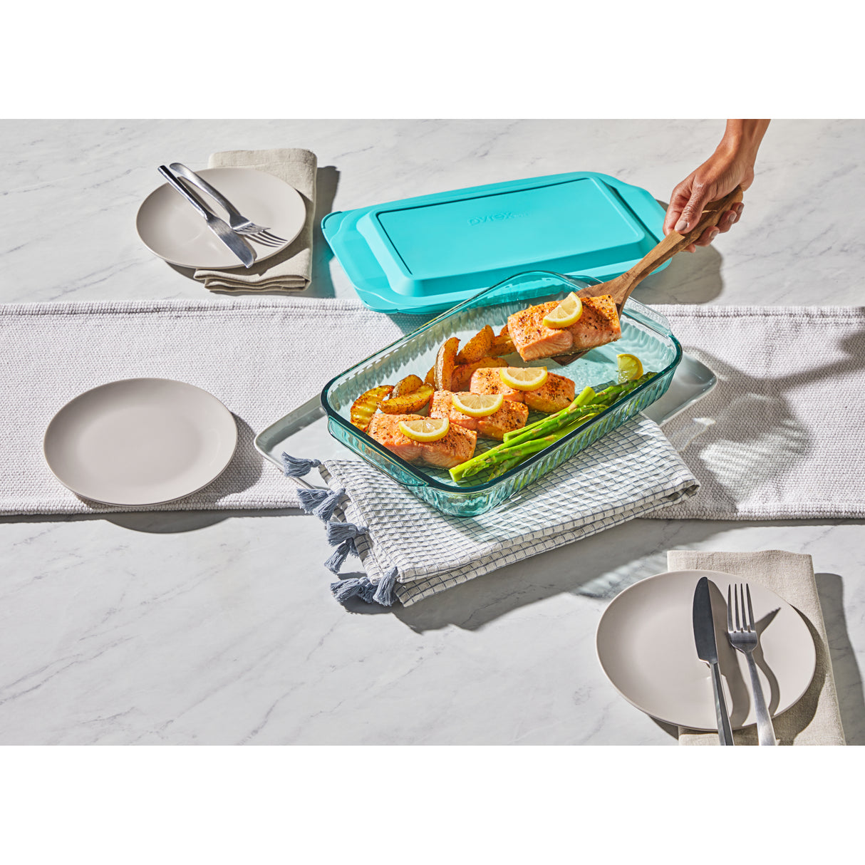 Glass dish with salmon and vegetables on a table with plates and cutlery.
