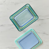 Set of glass food storage containers with blue and green gradient lids on a marble surface.