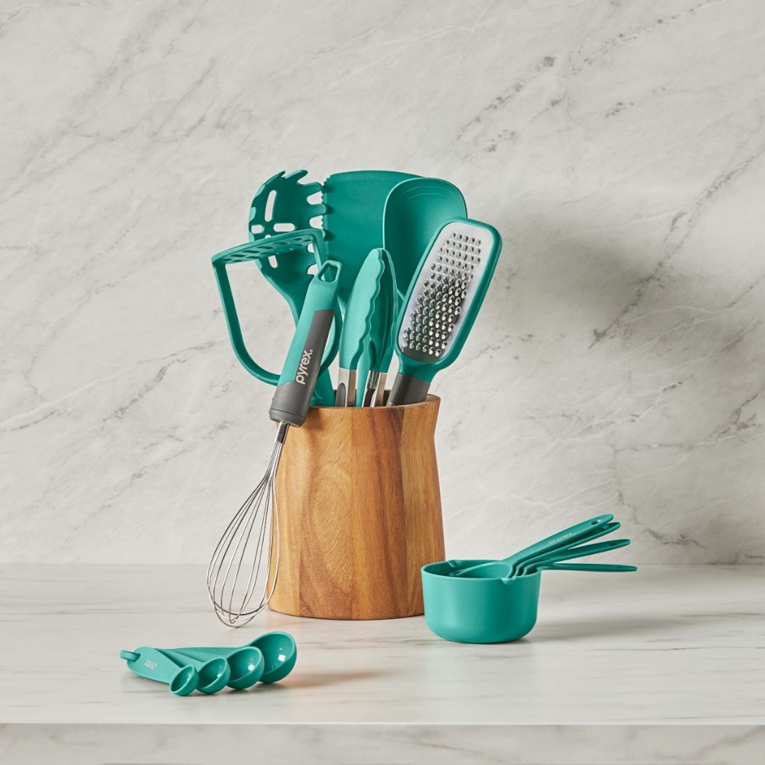 Teal kitchen utensils including a whisk, spatula, and measuring cups in a wooden holder on a marble countertop.