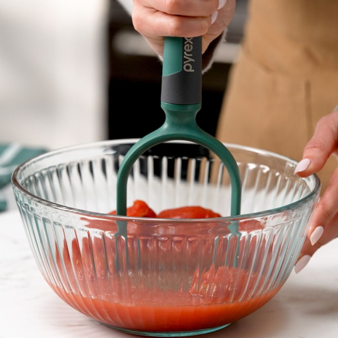 Person using a green potato masher in a glass bowl with red potatoes.