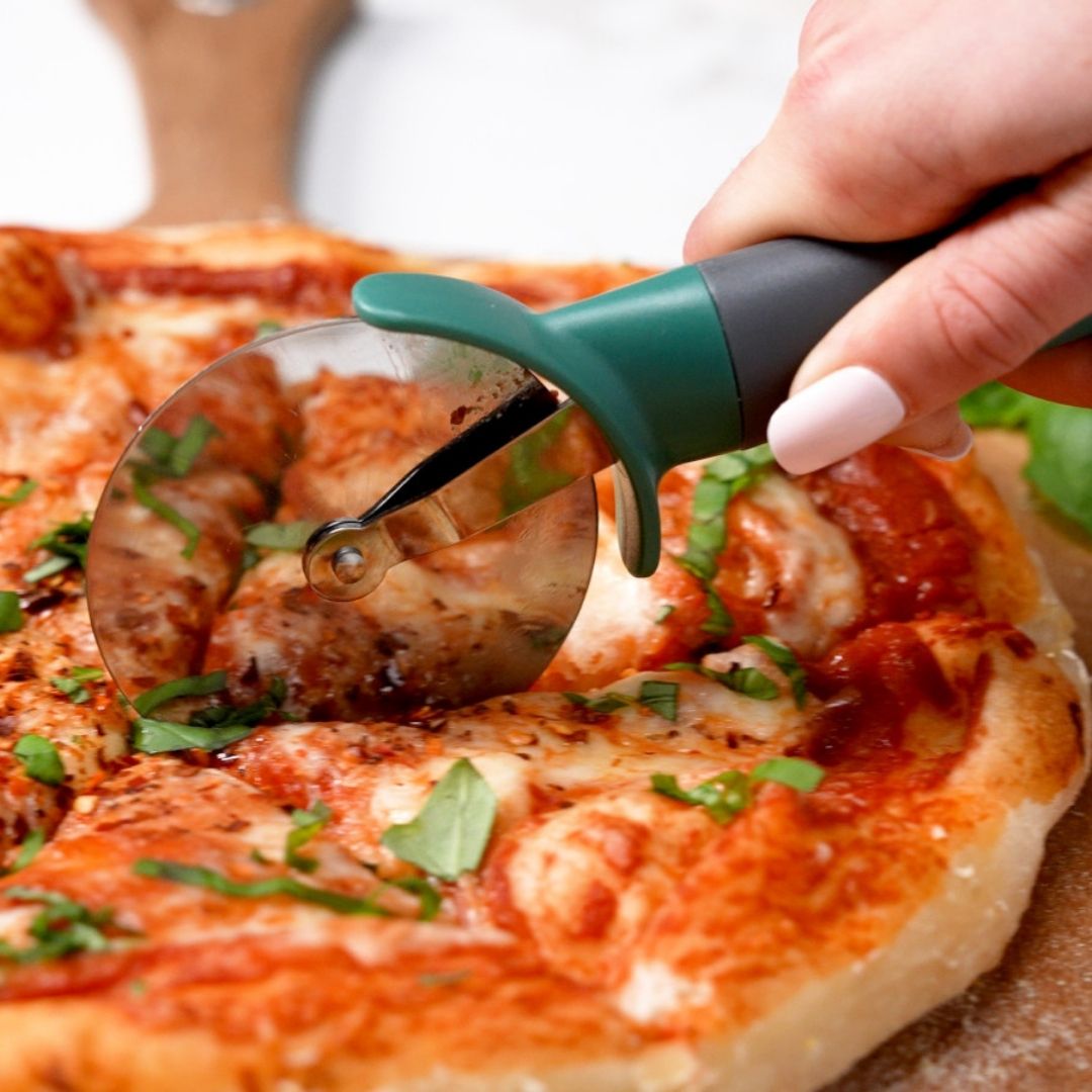 Hand using a pizza cutter to slice a pizza with visible toppings.