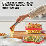 Person using tongs to serve pasta from a glass container with red lid, surrounded by other food containers on a kitchen counter.