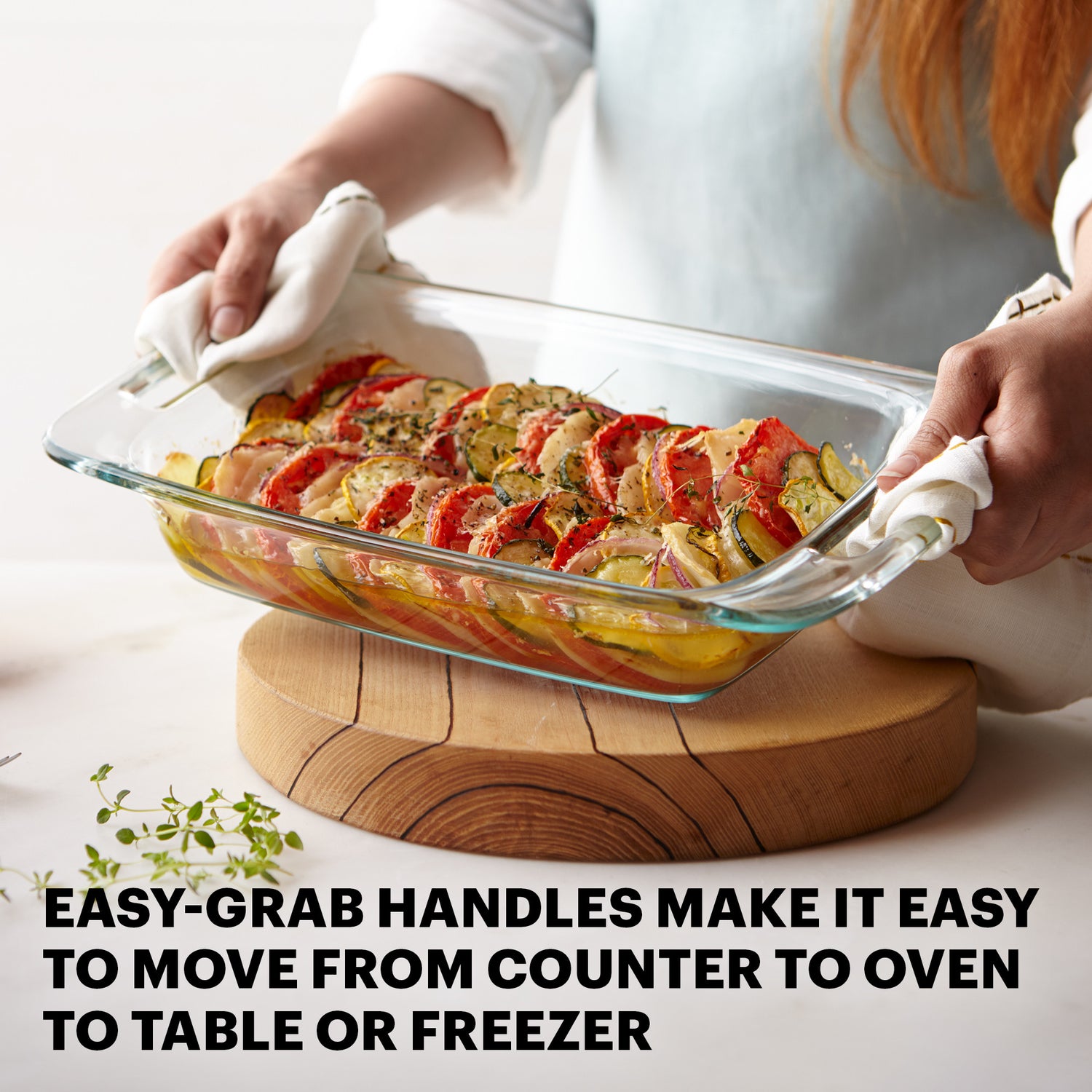 Glass baking dish with food held by a person on a wooden trivet, with text about easy-grab handles.