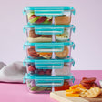 Stack of glass food containers with blue lids filled with various snacks on a pink background