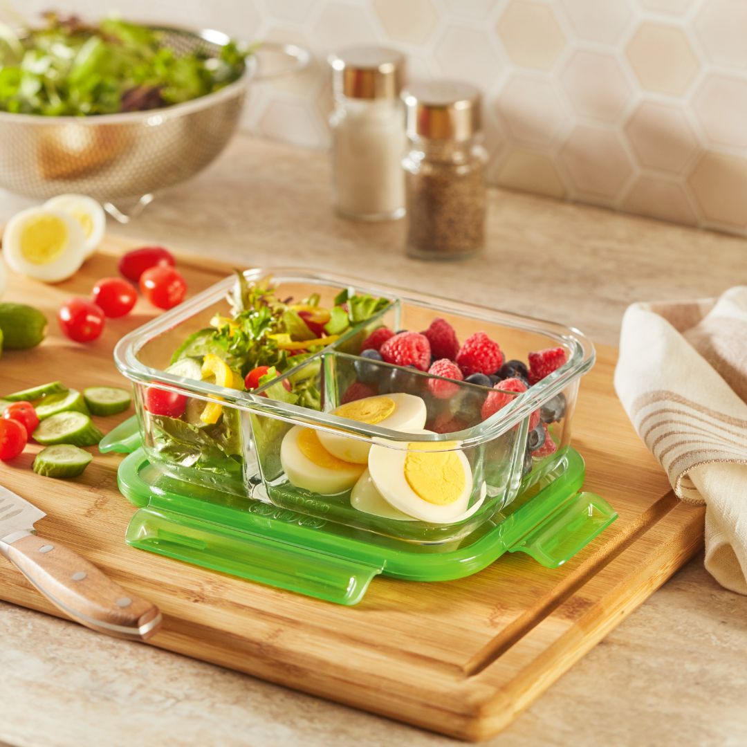 Glass food container with salad and hard-boiled eggs on a wooden cutting board with a kitchen background.