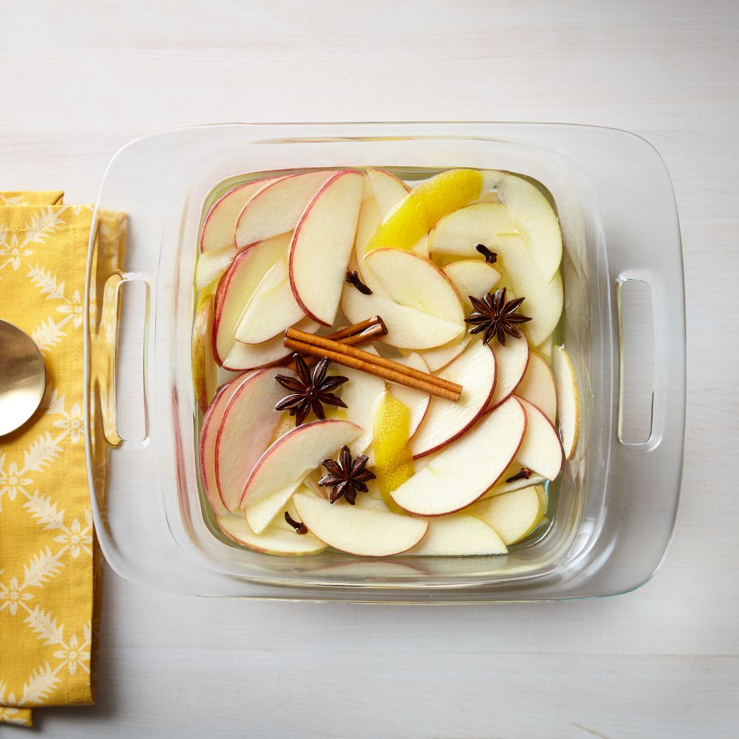 Glass dish with sliced apples, cinnamon sticks, and star anise on a white surface with a yellow cloth and spoon.
