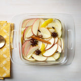 Glass dish with sliced apples, cinnamon sticks, and star anise on a white surface with a yellow cloth and spoon.