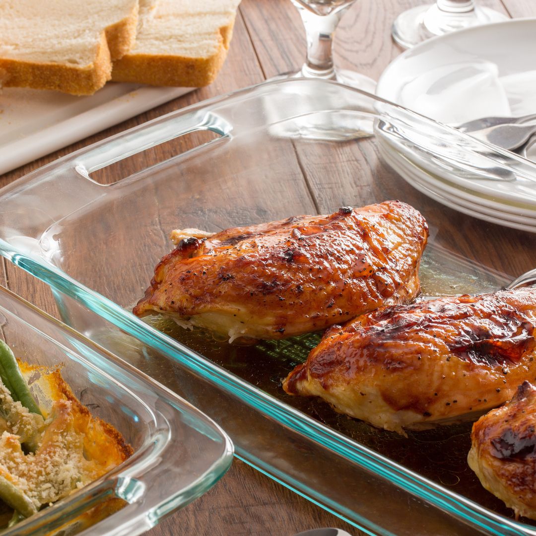 Glass dish with roasted chicken on a wooden table with bread and plates.