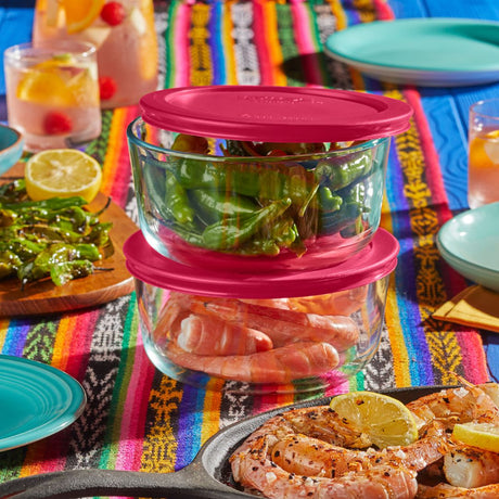 Stack of glass food containers with pink lids on a colorful tablecloth with food and drinks.
