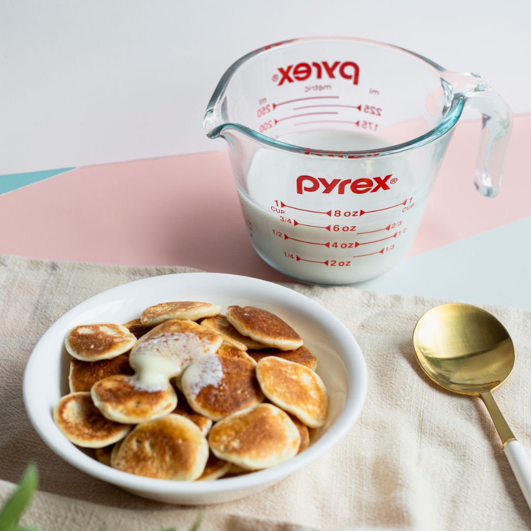Pyrex measuring cup with milk next to a bowl of pancakes on a light background