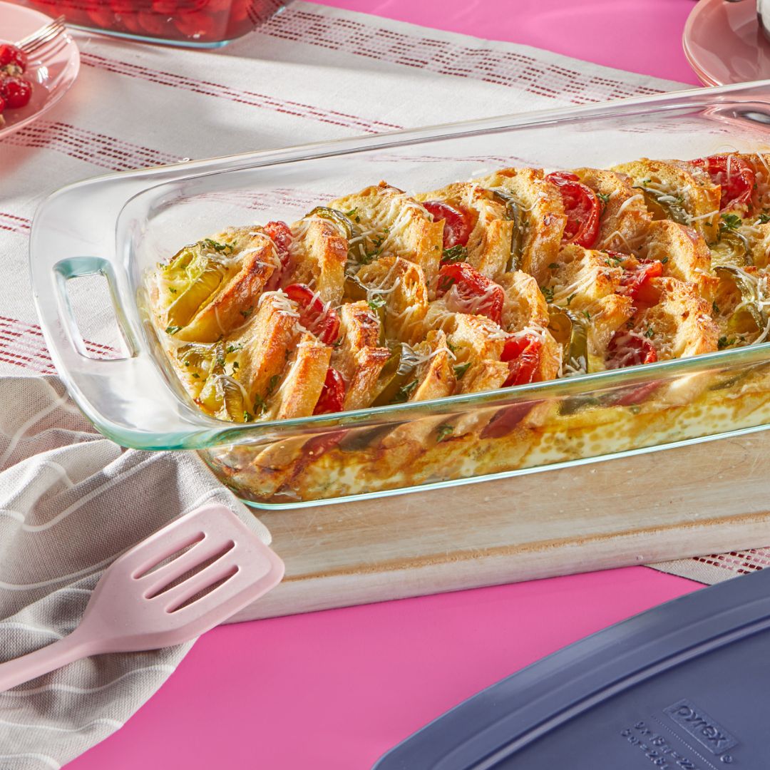 Glass dish with cheesy vegetable casserole on a pink surface with a pink spatula.