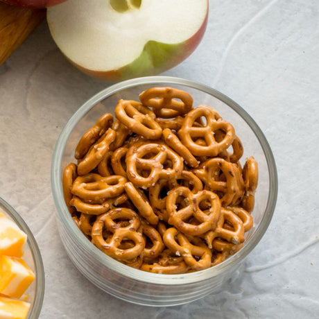 Clear glass bowl filled with pretzels on a light gray surface with an apple in the background.